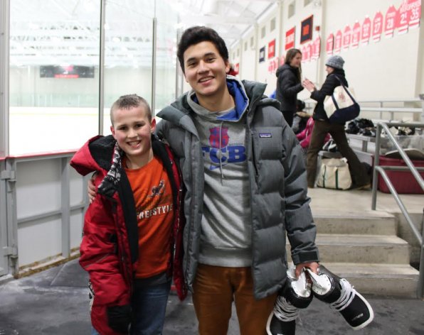 Friends Youth Mentoring Program mentor Paul Kigawa teaching his mentee, Stephen Johnson, how to ice skate – and how to pose for a picture in an ice arena – for the first time.