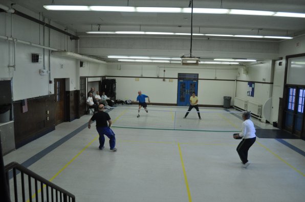 A crowd of anxious players await their turns as Larry Phillips fires a shot toward Jack LaFlamme during a pickleball showdown. The move indoors after a summer at Merrill Park has left just one court available at a time, which is bordered by obstacles such as waiting people and random equipment.