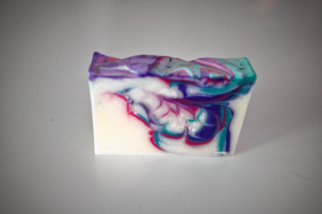 The finished bar of soap is primarily white with swirls of purple, teal and magenta.