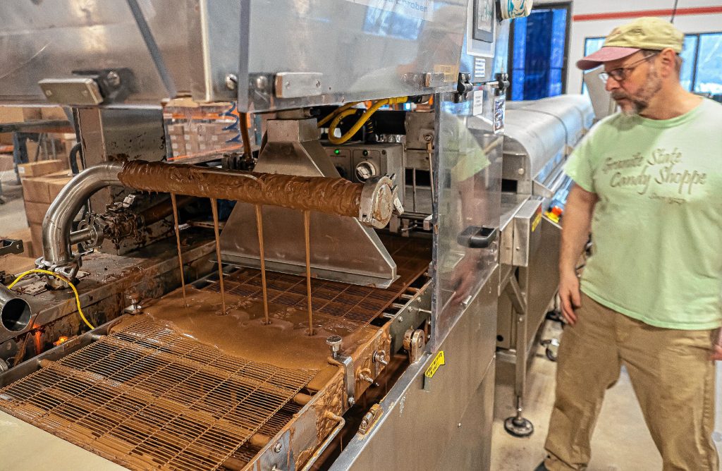 Jeff Bart, owner of the Granite State Candy Shoppe, stands by one of the enroving line machines where milk chocolate is dropped onto the top of a center filling at the new location for expanded production. GEOFF FORESTER