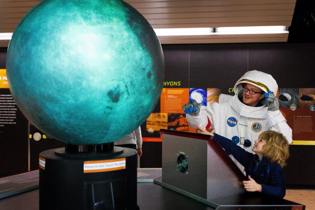 While wearing a replica of Alan ShepardÕs spacesuit, Jerrid Kenney helps 7-year-old Trase Boudreau of Beverly, Mass., use the OmniGlobe to look at the moon and different planets in the Looking at Earth, Looking Beyond exhibit at the McAuliffe-Shepard Discovery Center, Saturday, Nov. 22, 2014.  (ELIZABETH FRANTZ / Monitor staff) Visitors to the McAuliffe-Shepard Discovery Center last Saturday got to see an usual sight – a walking, talking astronaut. While the astronaut was really volunteer Jerrid Kenney of Manchester donning a replica of Alan Shepard’s spacesuit, young visitors and parents didn’t seem disappointed. The astronaut will be at the museum every Saturday from noon to 3:30 p.m. until Christmas. Above: Ada Wright, 3, gives a high-five to Kenney.Left: Kenney helps Trase Boudreau, 7, of Beverly, Mass., use the OmniGlobe.       ELIZABETH FRANTZ